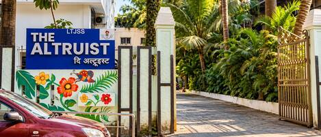 A gated entrance with a sign for 'The Altruist' hotel, surrounded by lush greenery and palm trees.