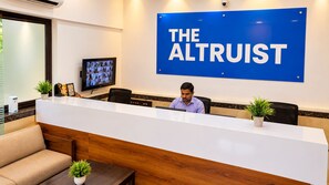A reception area with a large blue sign reading 'THE ALTRUIST,' a reception desk, a television mounted on the wall, and a person seated at the desk. - The Altruist Koparkhairane Hotel (Navi Mumbai)