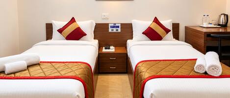 A hotel room with two beds, each with a red and gold bedspread, white linens, and a red and gold pillow. There is a wooden nightstand with a lamp, a small table with a kettle and water bottles, and a framed picture on the wall.