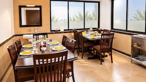 A dining area with a large table set for multiple people, a kitchen area with a stainless steel range, and large windows offering a view of greenery.