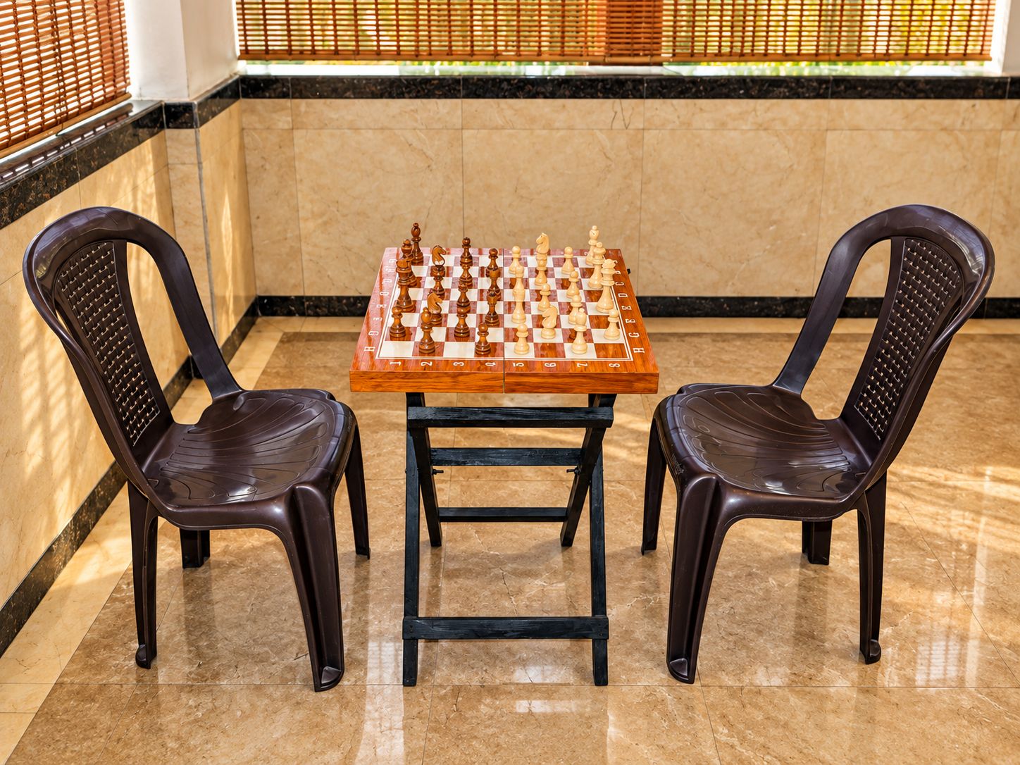 A chessboard set on a table with two chairs, in a room with a tiled floor and a window with blinds.