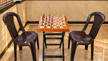 A chessboard set on a table with two chairs, in a room with a tiled floor and a window with blinds.