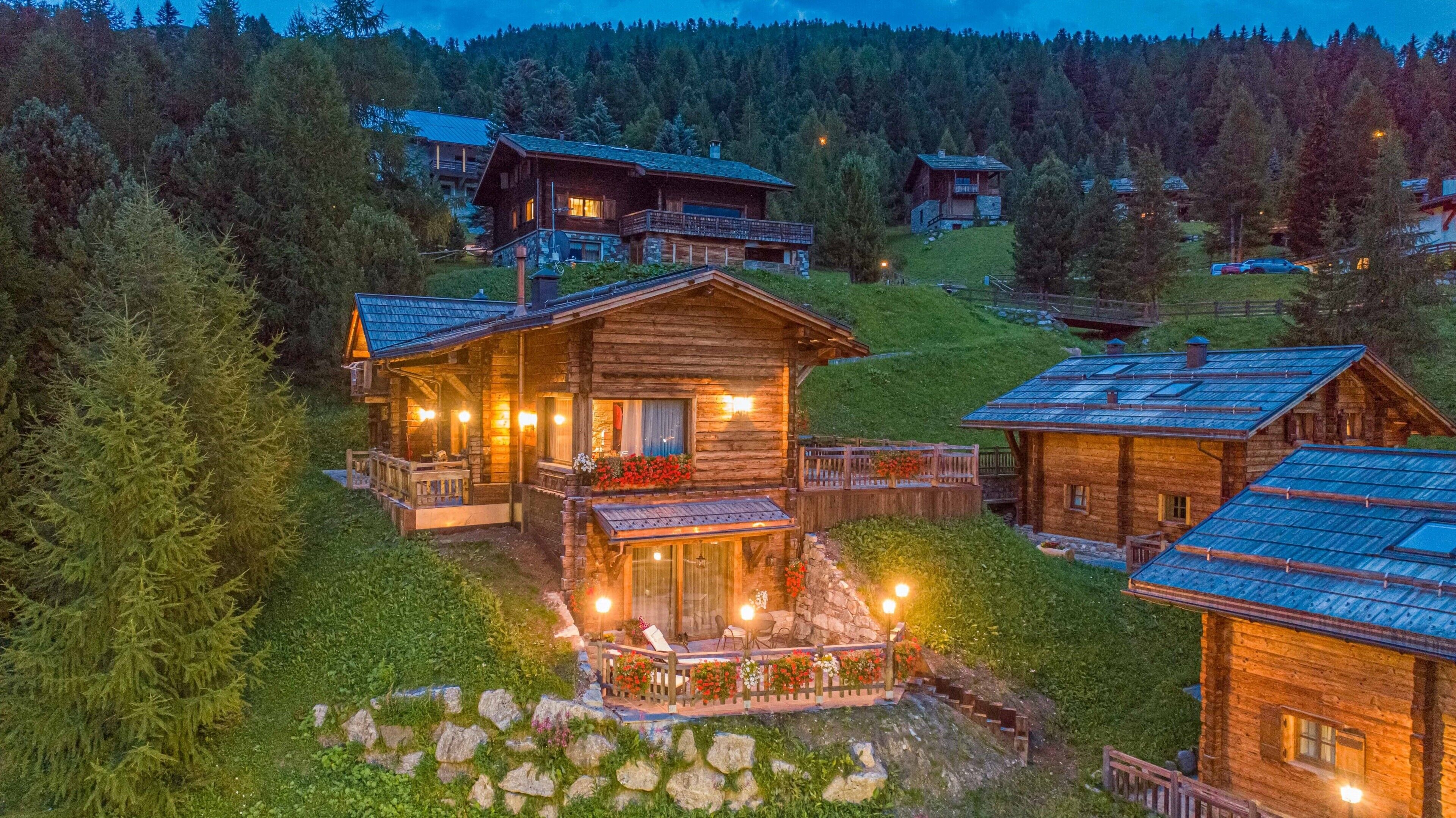 A wooden cabin with a balcony, surrounded by trees and a mountainous backdrop.
