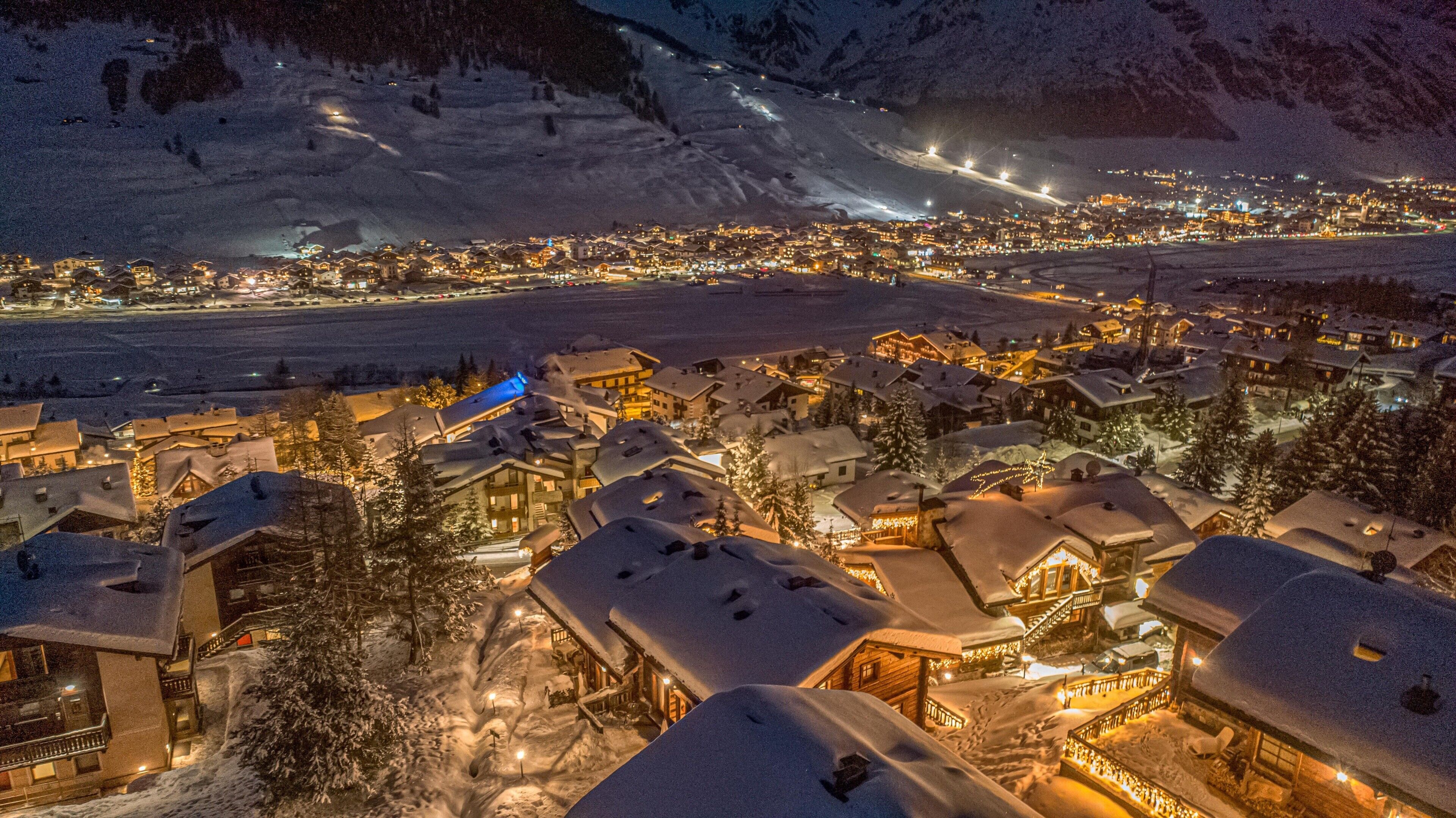 A snow-covered town with illuminated buildings and a ski slope in the background.