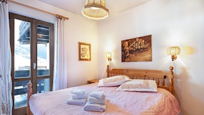 A bedroom with a bed, a wooden headboard, a window with curtains, a wall-mounted lamp, and a painting.