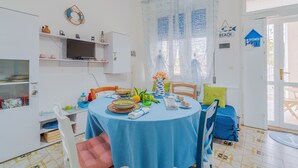 Uma área de refeições com uma mesa redonda, cadeiras e toalha de mesa azul. Há uma televisão em uma prateleira acima da mesa.
