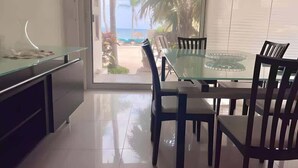 Une cuisine moderne avec une table et des chaises en verre, un îlot central et une vue sur la plage par une porte vitrée.