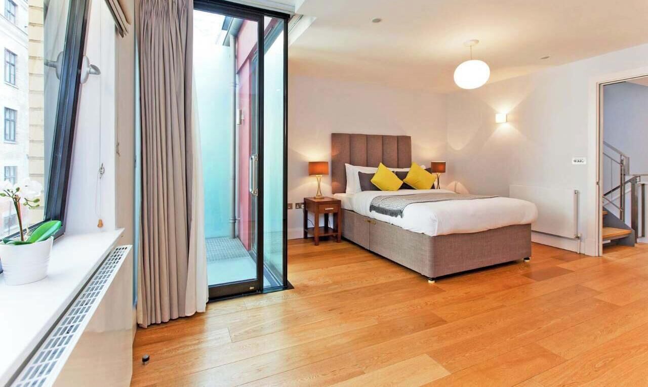 A modern hotel room with a large bed, a bedside table with a lamp, a sliding glass door leading to an outdoor area, and a staircase to the right.