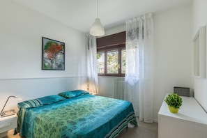 A bedroom with a bed, a desk, a potted plant, a window with curtains, and a wall-mounted picture.