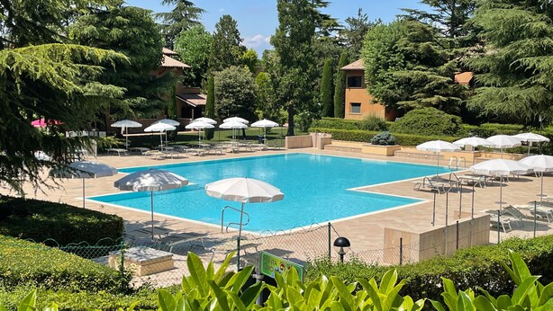 A swimming pool surrounded by lounge chairs and umbrellas, with a fence and greenery in the foreground. - Holiday Apartment "Casa Punta Grò" with Shared Pool, Terrace and Wi-Fi (Sirmione)