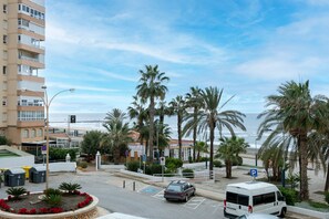 Una calle costera con un cartel de estacionamiento, palmeras, un edificio con balcones y vistas al mar.