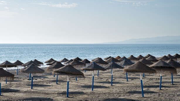Uma praia com fileiras de guarda-sóis de palha e um mar calmo ao fundo.