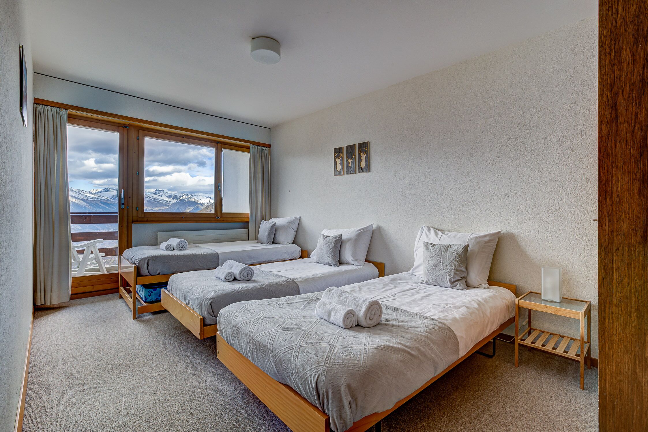 A bedroom with two beds, a window with a view of mountains, a bedside table with a lamp, and a wall-mounted picture.