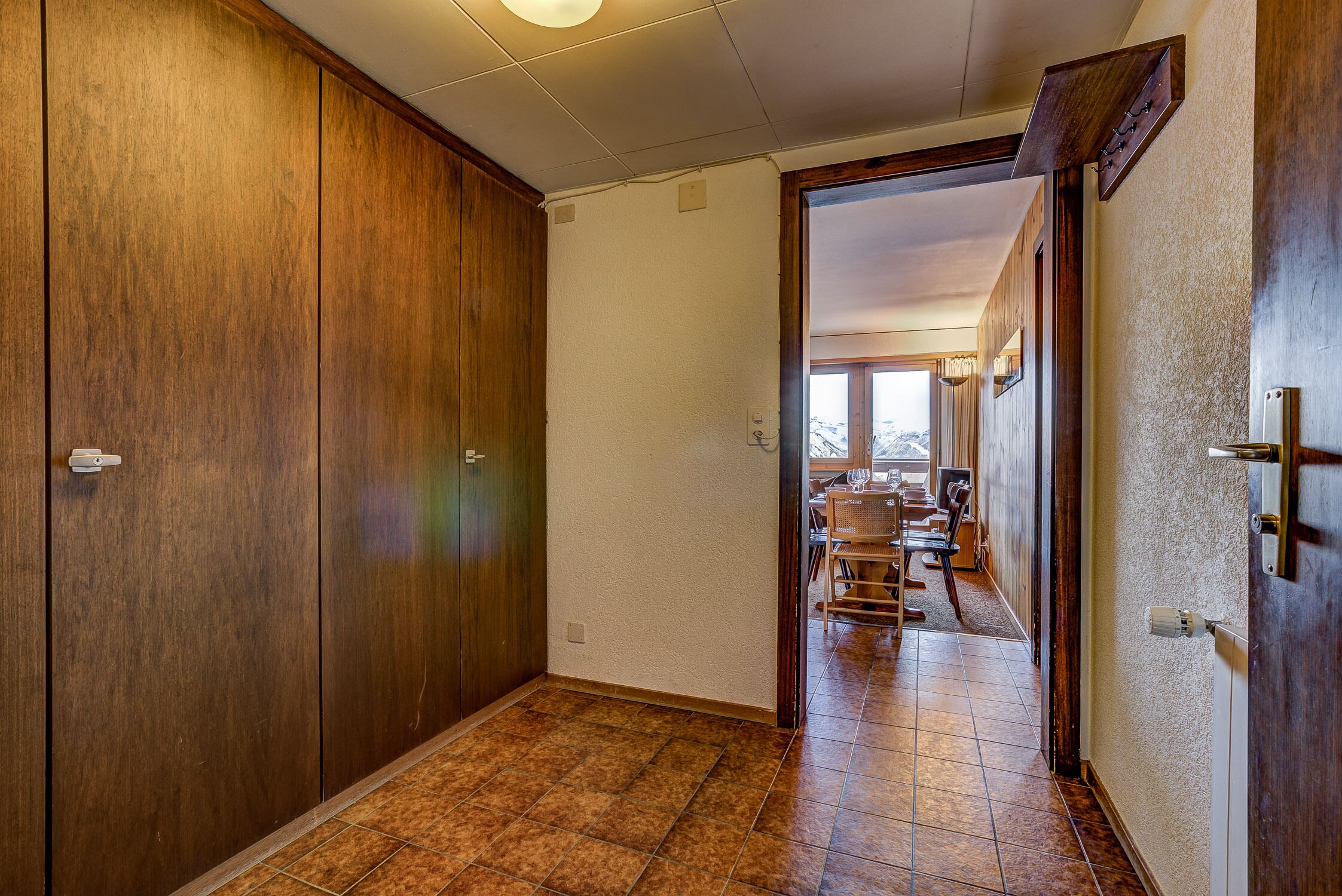 A hallway with wooden cabinets, a tiled floor, and a view into a dining area with chairs and a table.