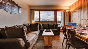 A cozy living room with a wooden dining table, leather sofa, and a view of mountains through large windows. - Appartement Mont Parrain 14 - La Tzoumaz, 4 Vallees (La Tzoumaz)