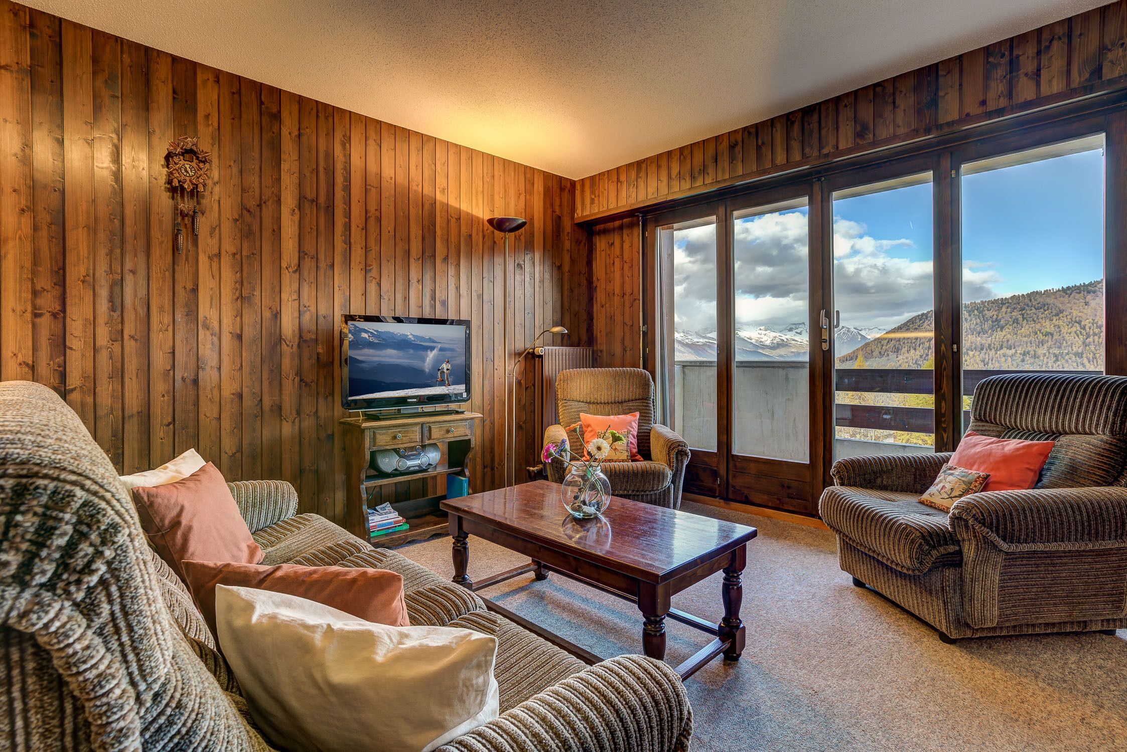 Ein gemütliches Wohnzimmer mit Holzwänden, einem Fernseher, einem Kamin und Bergblick durch große Fenster.