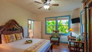 A bedroom with a wooden bed, a ceiling fan, a window with a view of greenery, and a television on the wall.