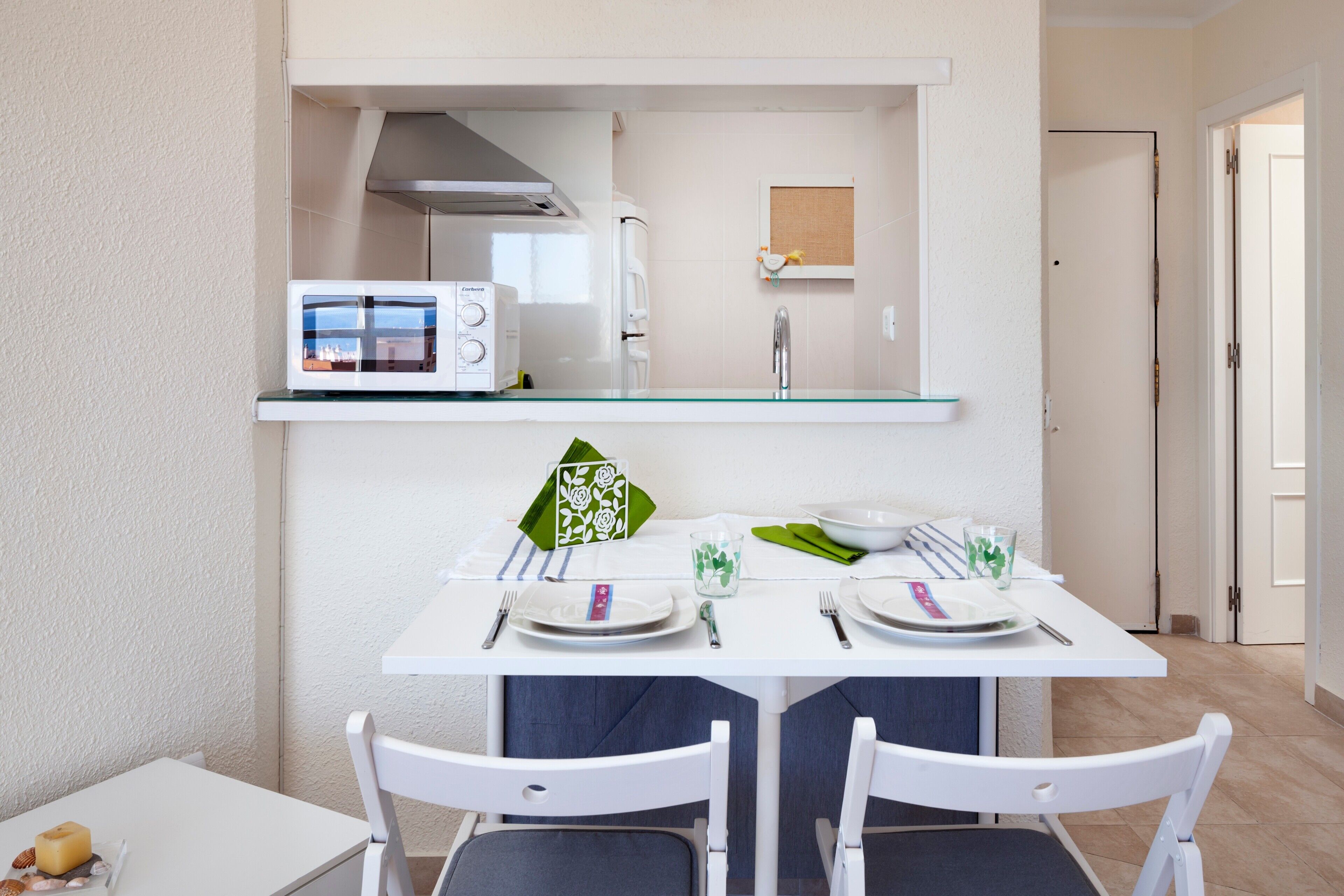 Une cuisine moderne équipée d’un four à micro-ondes, d’un évier et d’un plan de travail en verre. On y trouve une table à manger avec des chaises, ainsi qu’une petite table sur laquelle sont posés un chandelier et une boîte de bougies.