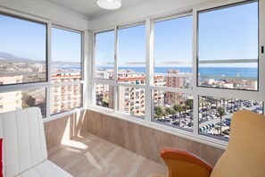 Een kamer met grote ramen, een witte bank en uitzicht op een stadsbeeld en de zee.