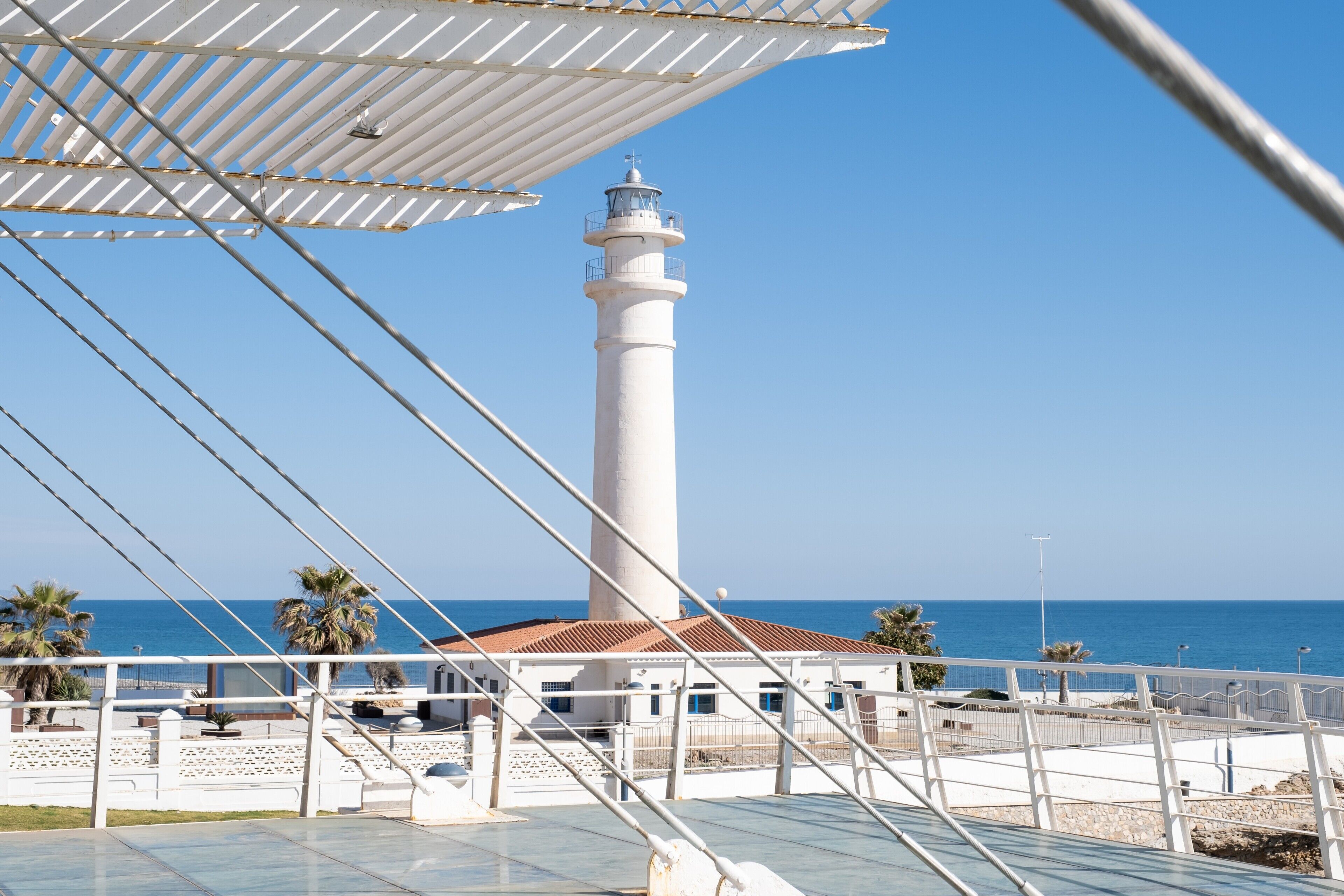 A lighthouse with a white tower and red roof, a white building with a red roof, and a clear blue sky.