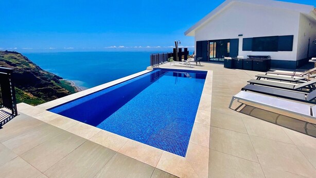 A modern pool with a clear view of the ocean, a white building with large windows, and a patio area with lounge chairs. - Holiday Home "Villa Camacho xxvii" with Sea View, Pool & Wi-Fi (Arco da Calheta)