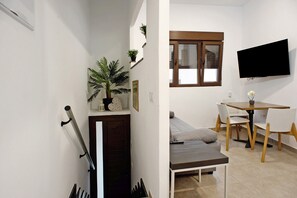 Una habitación pequeña y moderna con sofá, una mesa de comedor con sillas, televisión montada en la pared y una escalera con una planta en la barandilla.