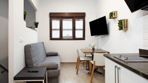 A compact living space with a sofa, a dining table, chairs, a TV mounted on the wall, and a window with blinds. - Vacation 'Jardin Cardona' with Wi-Fi (Arucas)