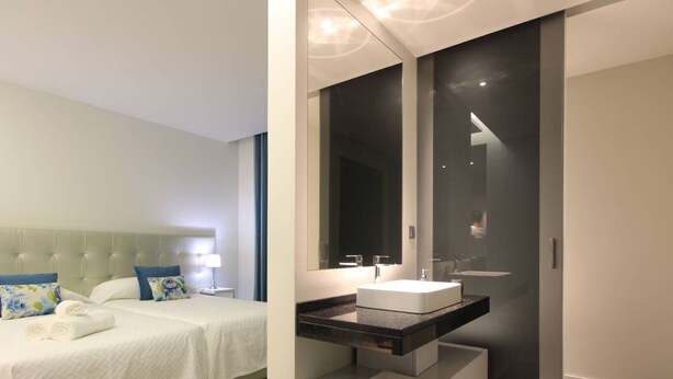 Een moderne hotelkamer met een bed, een grote spiegel, een witte wastafel en een inbouwkleerkast.