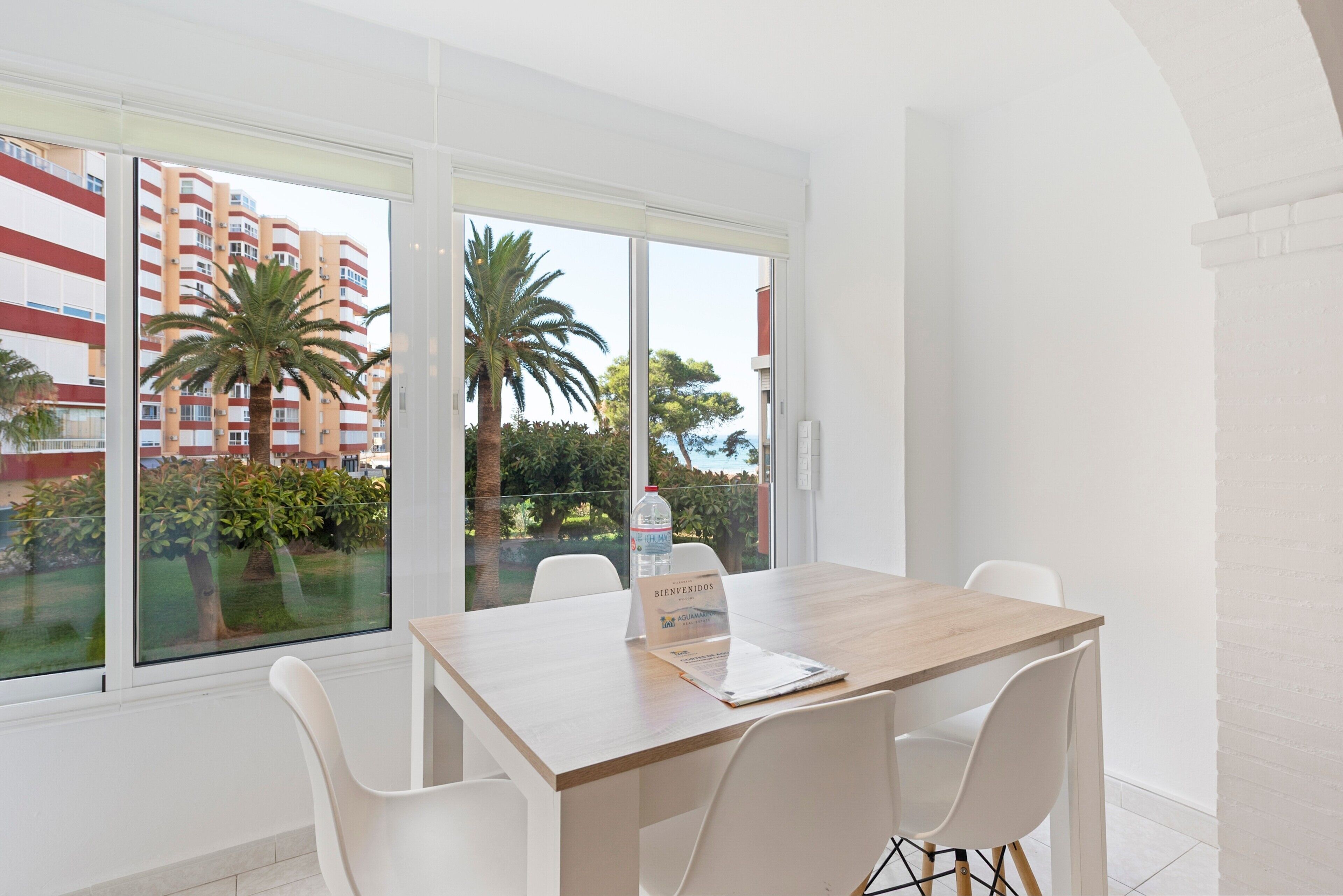 Una habitación moderna con una mesa de madera, sillas blancas y ventanas amplias con vistas a un edificio y palmeras.