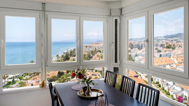 A dining area with a table set for four, large windows, and a view of the sea and cityscape. - Apartment "Vistas Playa Torrox A73" with Sea View, Shared Pool and Wi-Fi (Torrox)
