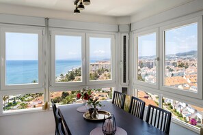 A dining area with a table set for four, large windows, and a view of the sea and cityscape. - Apartment "Vistas Playa Torrox A73" with Sea View, Shared Pool and Wi-Fi (Torrox)