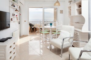 A room with a TV, white furniture, a table with chairs, and a view of the sea.