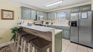 Una cocina con isla central, taburetes de bar, refrigerador, microondas y cafetera.