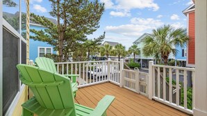 Una terraza de madera con sillas Adirondack verdes, barandilla blanca y vistas a casas coloridas y palmeras.