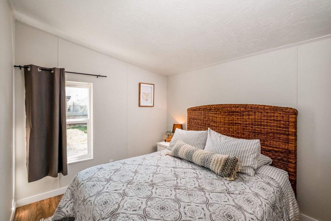 A bedroom with a bed, a window with curtains, a bedside table with a lamp, and a framed picture on the wall.