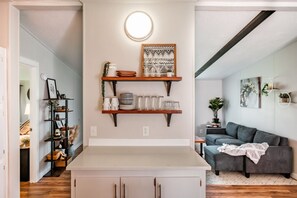 Une cuisine moderne avec un plan de travail blanc, des étagères en bois et un canapé gris dans l’espace de vie adjacent.