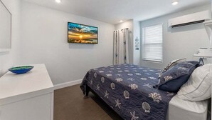 A bedroom with a bed, a desk, a window with blinds, an air conditioner, and a painting on the wall. - Beautifully Renovated Beach Block Condo! (Wildwood)