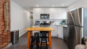 A compact kitchen with white cabinets, a stainless steel refrigerator, a wooden table with stools, and a brick accent wall. - Beautifully Renovated Beach Block Condo! (Wildwood)