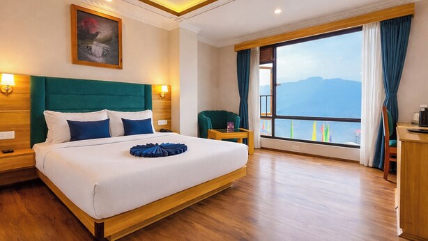 A hotel room with a large bed, a view of mountains, a wooden floor, and a nightstand with a lamp. - Yashshree Sikkim Blossom 600 Meters From Mg Marg (Gangtok)