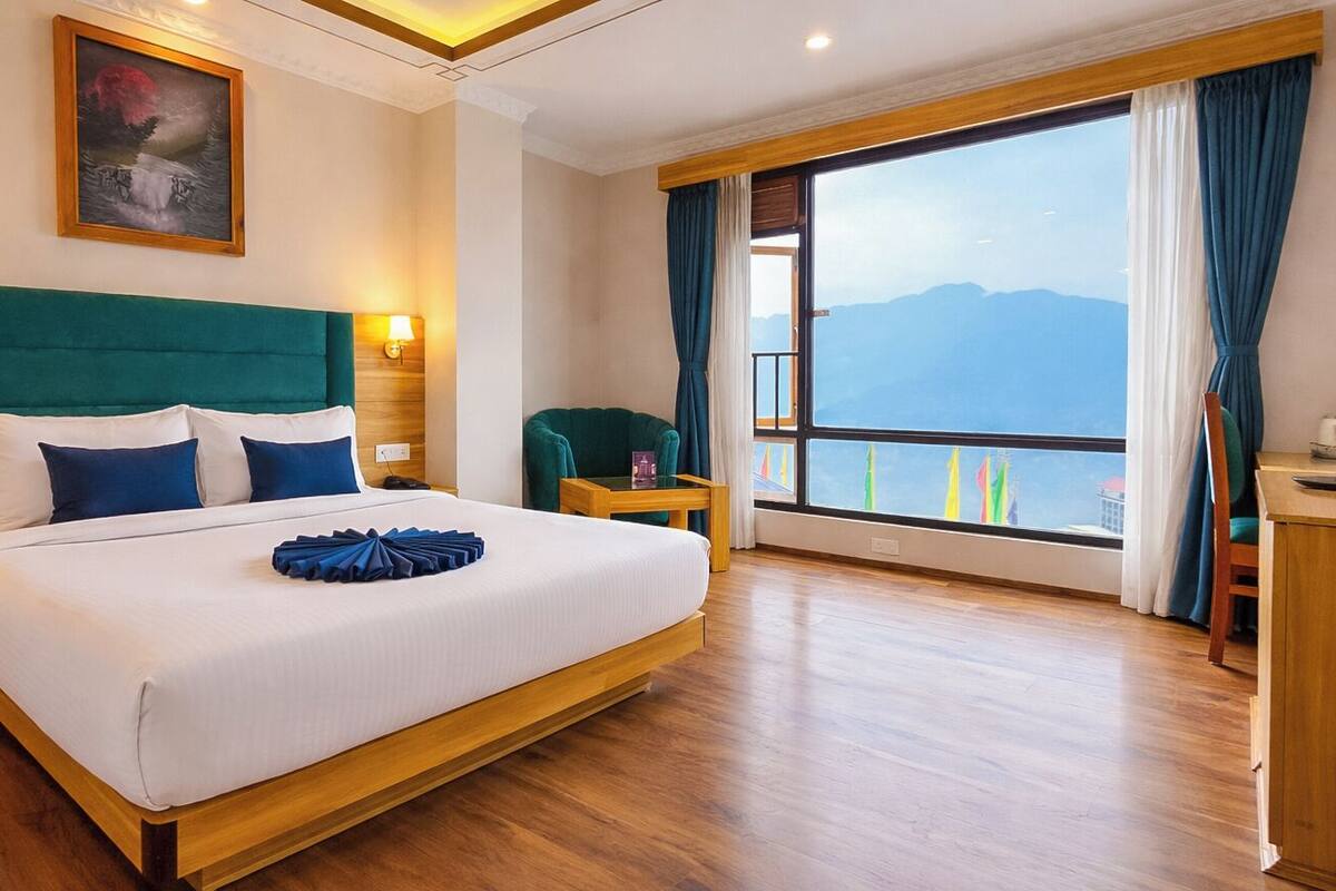 A hotel room with a large bed, a view of mountains, a wooden floor, and a nightstand with a lamp.