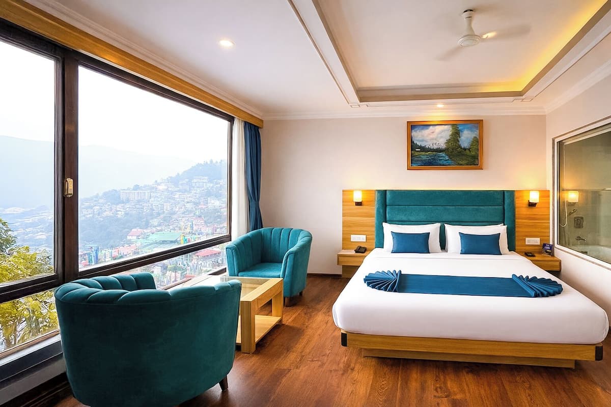 A hotel room with a large bed, a view of a cityscape, a painting on the wall, and a ceiling fan.