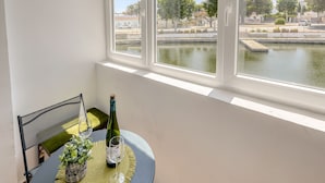 Un balcón con una mesa dispuesta para dos personas, una botella de vino y una copa. Se aprecia una vista de un canal y árboles a través de la ventana.