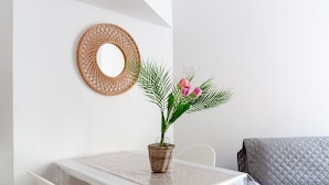 Et spiseområde med et hvidt bord, stole, en flettet kurv med en plante og et rundt spejl på væggen.