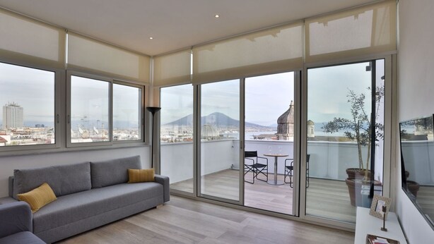 Una sala moderna con un sofá gris, una mesita y un balcón con vistas a un paisaje urbano y montañas.