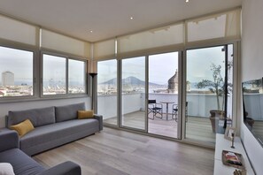 Un salon moderne avec un canapé gris, une petite table et un balcon offrant une vue sur le paysage urbain et les montagnes.