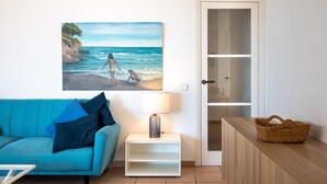 A living room with a blue sofa, a painting of a beach scene, a white side table with a lamp, and a wooden cabinet. - Seafront apartment with pool and parking in Torredembarra (Torredembarra)