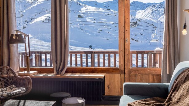 A cozy room with a large window overlooking a snowy mountain landscape, wooden beams, a wicker chair, and a radiator.
