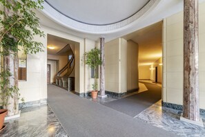 Un couloir au sol de marbre, avec une colonne centrale, un escalier et des plantes en pot.