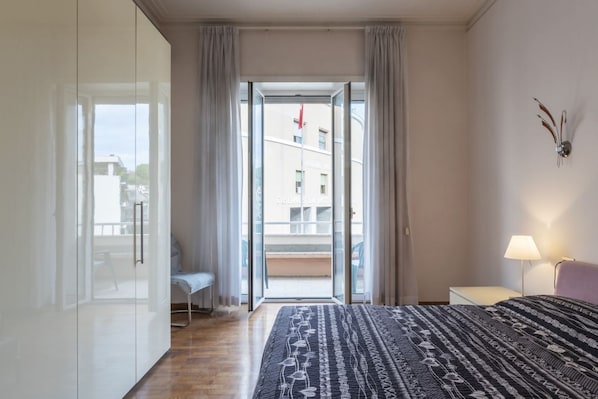 A bedroom with a bed, a bedside table with a lamp, a chair, a sliding glass door to a balcony, and a view of buildings outside.