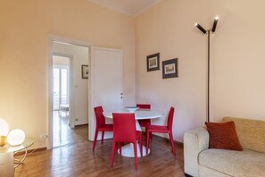 A room with a round dining table, four red chairs, a sofa, a floor lamp, and two framed pictures on the wall.